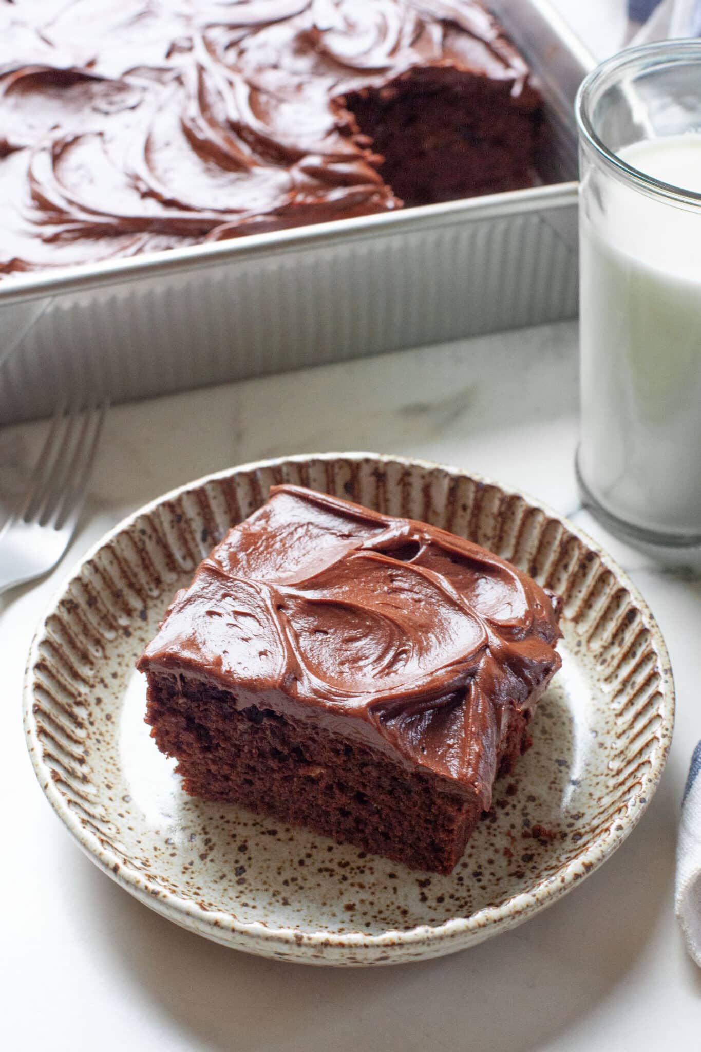 A slice of chocolate mayonnaise cake on a plate, with the remaining cake in a pan behind it.