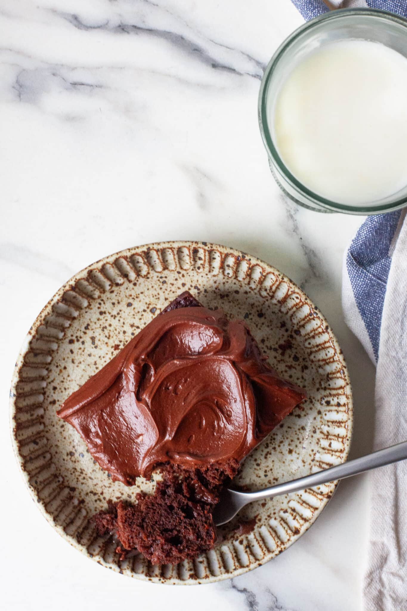 A slice of chocolate mayonnaise cake on a plate, with a piece of the cake on a fork. Next to the cake is a glass of milk.