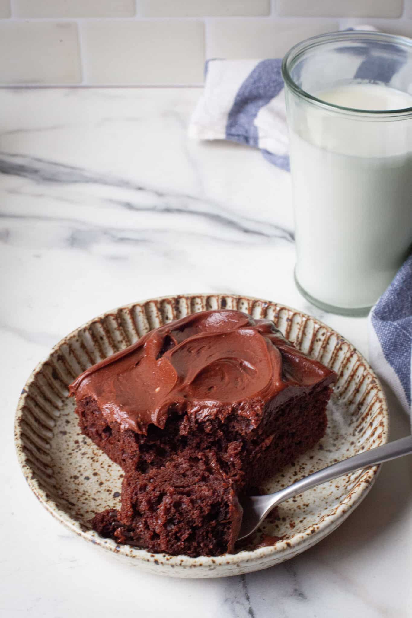 A slice of chocolate mayonnaise cake on a plate with a piece of cake on a fork. There is a glass of milk behind the plate.